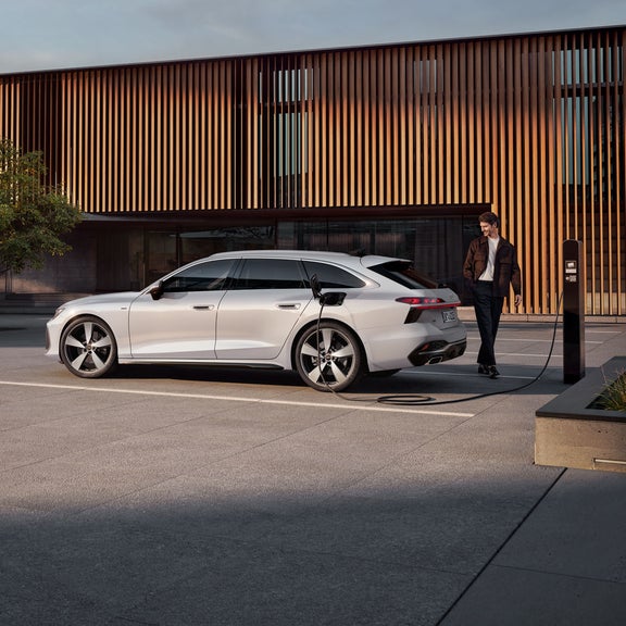 Der A6 Avant e-hybrid steht seitlich vor dem Eingangsbereich eines großen Gebäudes und wird über ein Stromkabel geladen. Am Heck des Fahrzeugs steht ein Mann mittleren Alters.