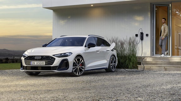 Der A5 Avant e-hybrid ist an eine Ladestation an einem Wohnhaus angeschlossen. Im Hintergrund verlässt ein Mann das Gebäude und blickt auf das Fahrzeug.