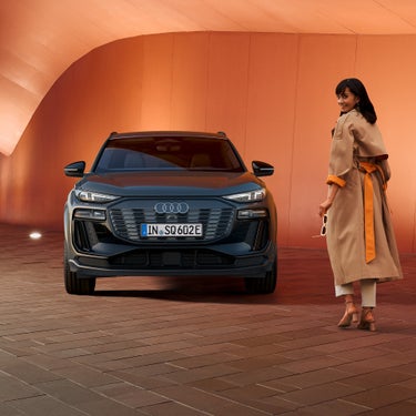 Front eines grauen Audi SQ6 SUV e-tron, rechts läuft Frau im Trenchcoat mit Sonnenbrille und lächelt. Im Hintergrund orangene Wand.