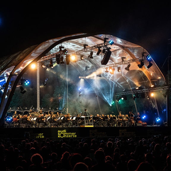 Orchester spielt nachts unter einer beleuchteten Freiluftbühne vor Publikum.