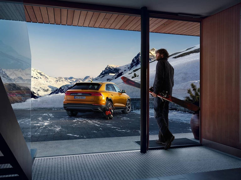 Someone walks out of the glass door of a hut in the mountains, an orange Audi is parked in front of the door.