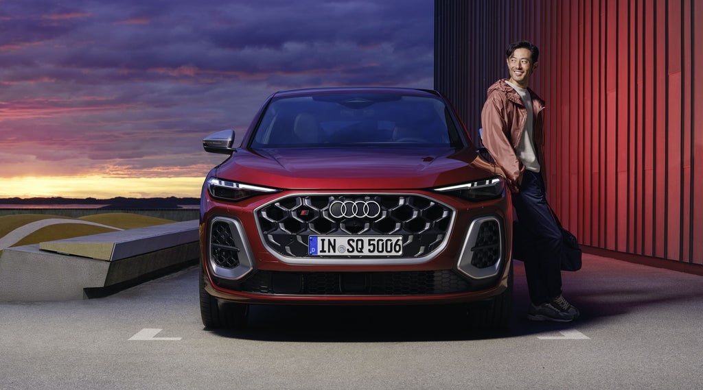 Der rote SQ5 Sportback steht auf einem Parkplatz parallel zu einem Hafen. Ein junger Mann lehnt sich an das Fahrzeug.