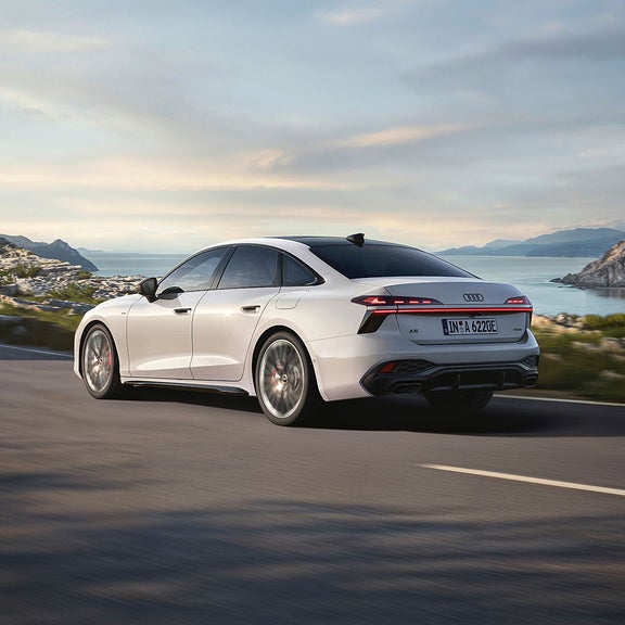 A white Audi A6 Saloon e-hybrid drives along a coastal road, showcasing its modern design against a scenic backdrop of mountains and blue water.