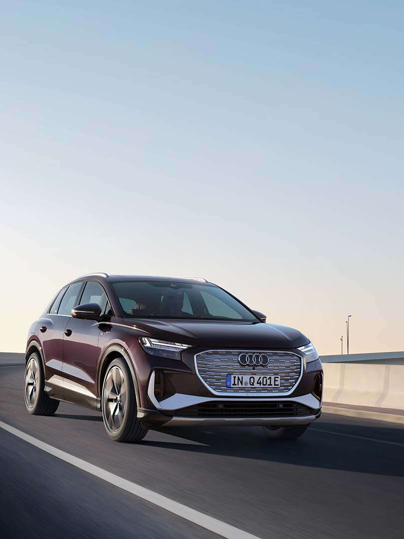 Violetter Audi Q4 e-tron fährt bei Tageslicht und blauem Himmel auf städtischer Straße mit modernen Hochhäusern im Hintergrund.
