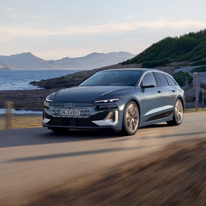 Der S6 Avant e-tron aus schräger Frontansicht, wie er eine Straße an der Küste entlangfährt. Im Hintergrund sind Berge zu sehen.