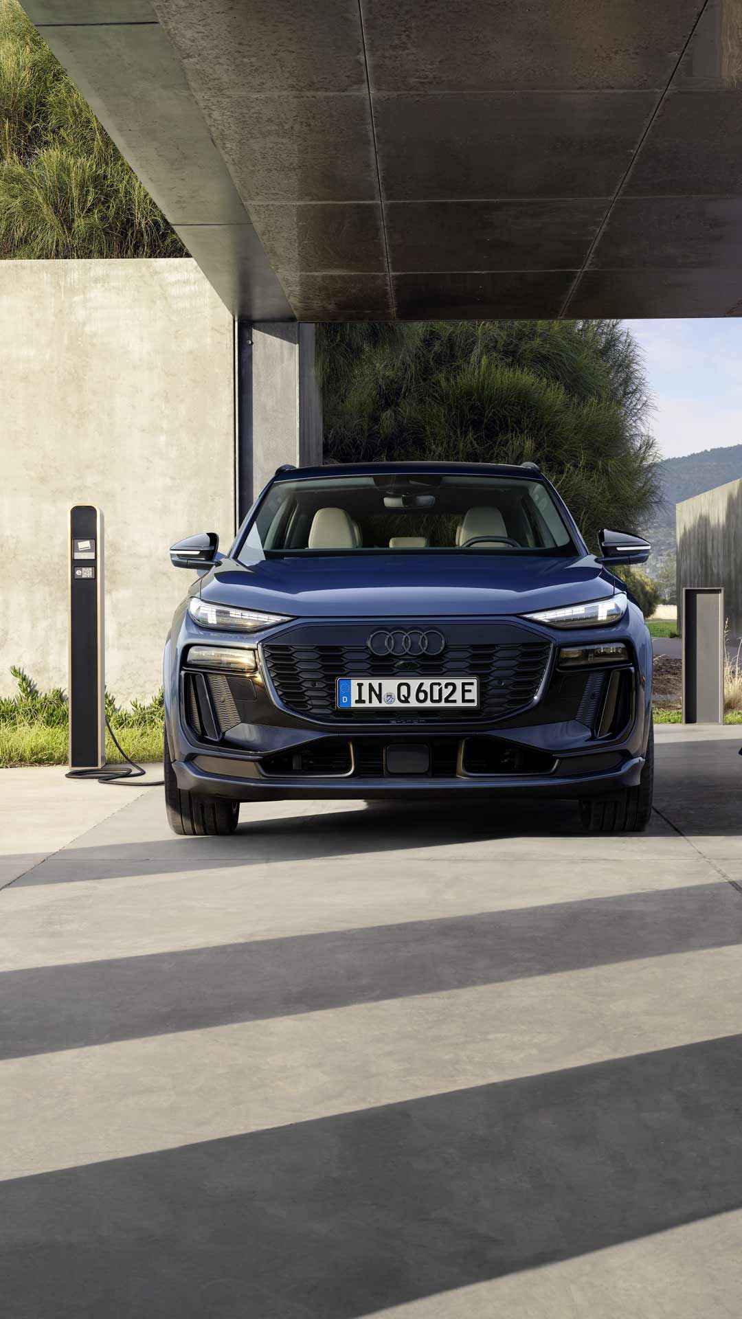 Frau mit Smartphone geht vor einem ladenden, blauen Audi Q6 SUV e-tron unter offenem Carport vorbei, im Hintergrund Naturkulisse vorbei.