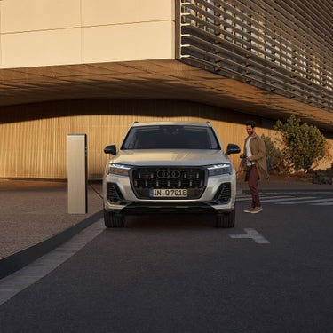 Frontansicht eines Audi Q7 SUV TFSI e an Ladesäule, Mann in beigem Mantel hält Ladekabel, moderne helle Gebäudefassade im Hintergrund.