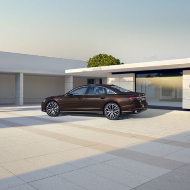 Seitliche Außenansicht eines braunen Audi A8 vor modernem, hell-beigen Haus bei Tageslicht.