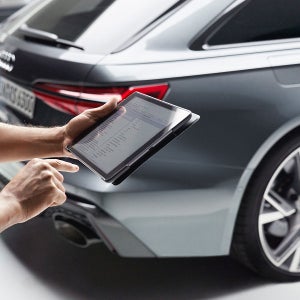 A person uses a tablet near a sleek, silver car, possibly for diagnostic purposes. The scene suggests modern technology and automotive expertise.