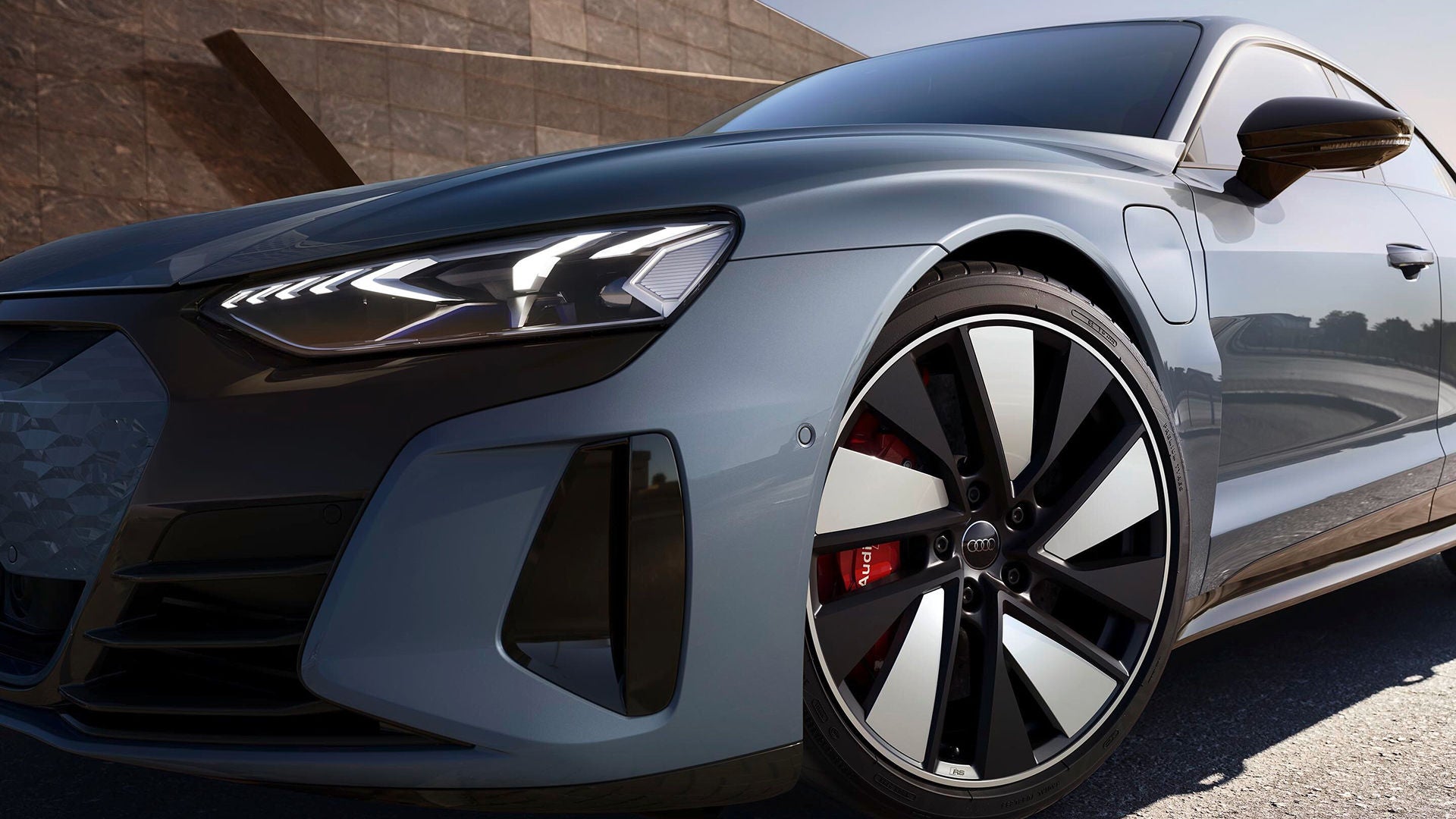 Close-up of a sleek, grey Audi car emphasising its front left headlight and modern alloy wheel. The polished surface reflects a sunny sky, conveying luxury and innovation.