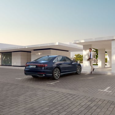 Mann in hellem Anzug läuft zu einem geparktem Audi A8 auf gepflastertem Platz, im Hintergrund ein helles Gebäude bei Tageslicht.