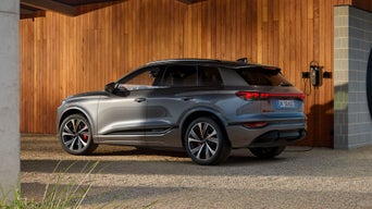 Seitenprofil des SQ6 SUV e-tron, welcher vor einer Holzhausfassade steht und an eine an der Hauswand montierte Ladestation angeschlossen ist.