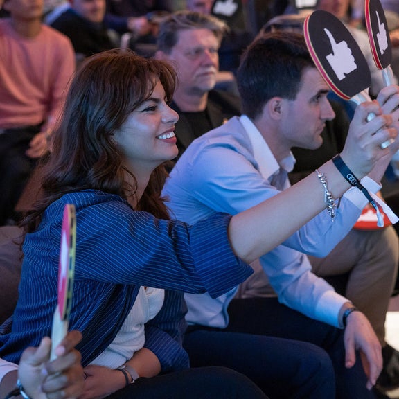 Spectators sit in the audience and hold voting signs with thumbs up or down. They look towards the front of the stage.