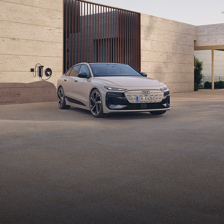 Weißer Audi steht vor moderner Hauswand, an der eine Ladestation installiert ist.