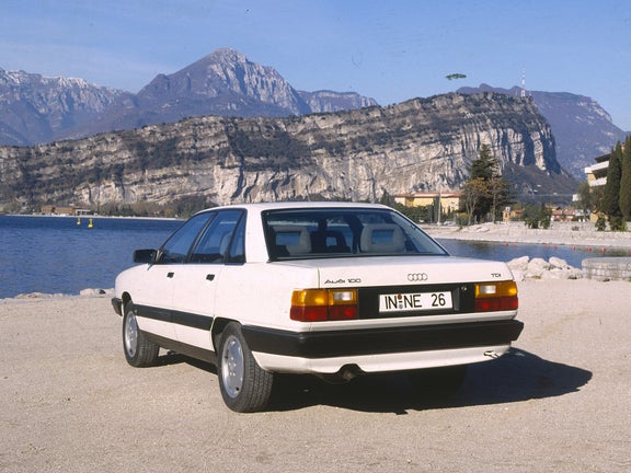 A white Audi 100 TDI in the rear view in front of a mountain backdrop with a mountain lake