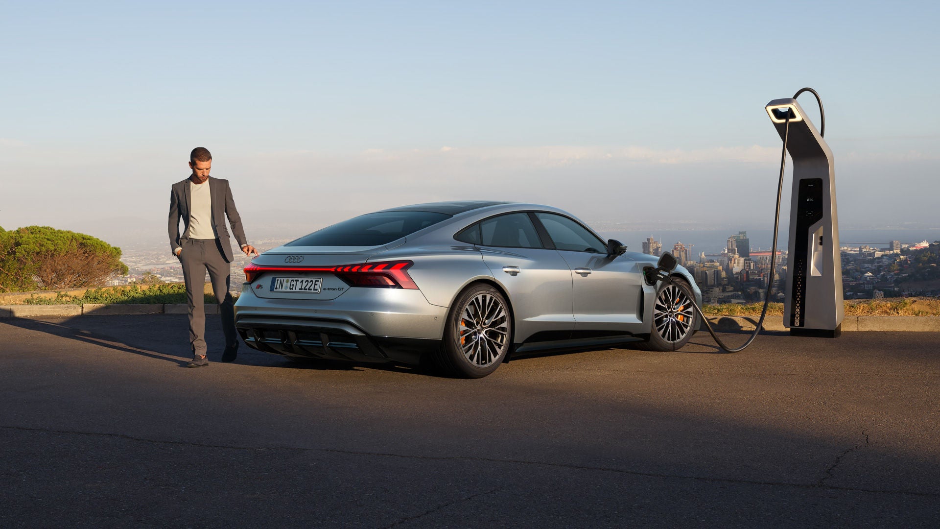 Grauer Audi S e-tron GT parkt an Ladesäule. Neben dem Auto steht ein Mann. Im Hintergrund ist eine Stadtsilhouette zu sehen.