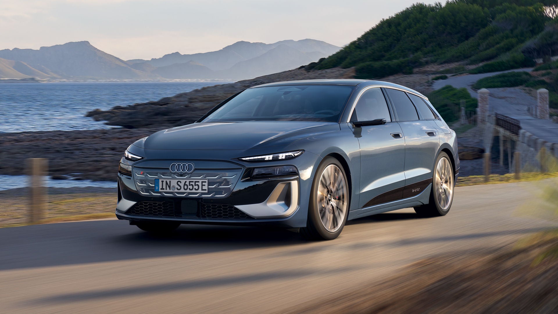Der S6 Avant e-tron aus schräger Frontansicht, wie er eine Straße an der Küste entlangfährt. Im Hintergrund sind Berge zu sehen.
