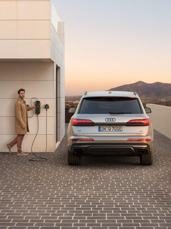 Rückansicht eines silbernen Audi Q7 SUV TFSI e, Mann an Wallbox, Frau im hellen Anzug, im Hintergrund Berge bei Sonnenuntergang.