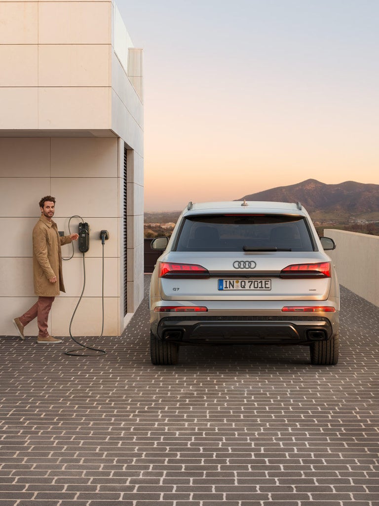 Rückansicht eines silbernen Audi Q7 SUV TFSI e, Mann an Wallbox, Frau im hellen Anzug, im Hintergrund Berge bei Sonnenuntergang.