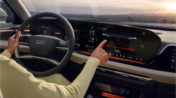 Fahrerin im gelben Outfit bedient großes Touchdisplay im Audi, Blick auf Digitalanzeigen und Landschaft bei Sonnenuntergang.