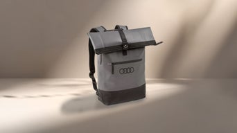 Sac à dos Audi gris avec fermeture à enroulement, poche avant et logo Audi, en plan large sur un fond clair et neutre, avec des effets d'ombre et de lumière doux.