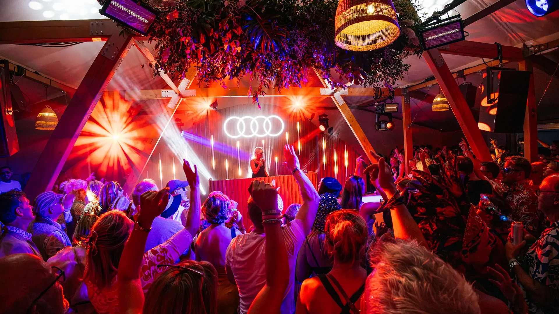 A large nighttime crowd gathers beneath bunting and the illuminated Audi rings outside the festival pavilion, with performers and a car visible amid the celebration. A large nighttime crowd gathers beneath bunting and the illuminated Audi rings outside the festival pavilion, with performers and a car visible amid the celebration.