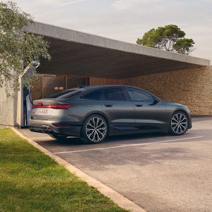 Grey Audi parked under a modern concrete carport with stone walls, surrounded by greenery on a sunny day, conveying luxury and elegance.