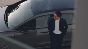 Aufnahme eines Audi A6 e-trons von schräg oben durch ein Fenster. Im Vordergrund unscharf der graue Fensterrahmen. Vor dem Audi steht ein Mann in blauem Anzug und mit Brille und blickt nach links.