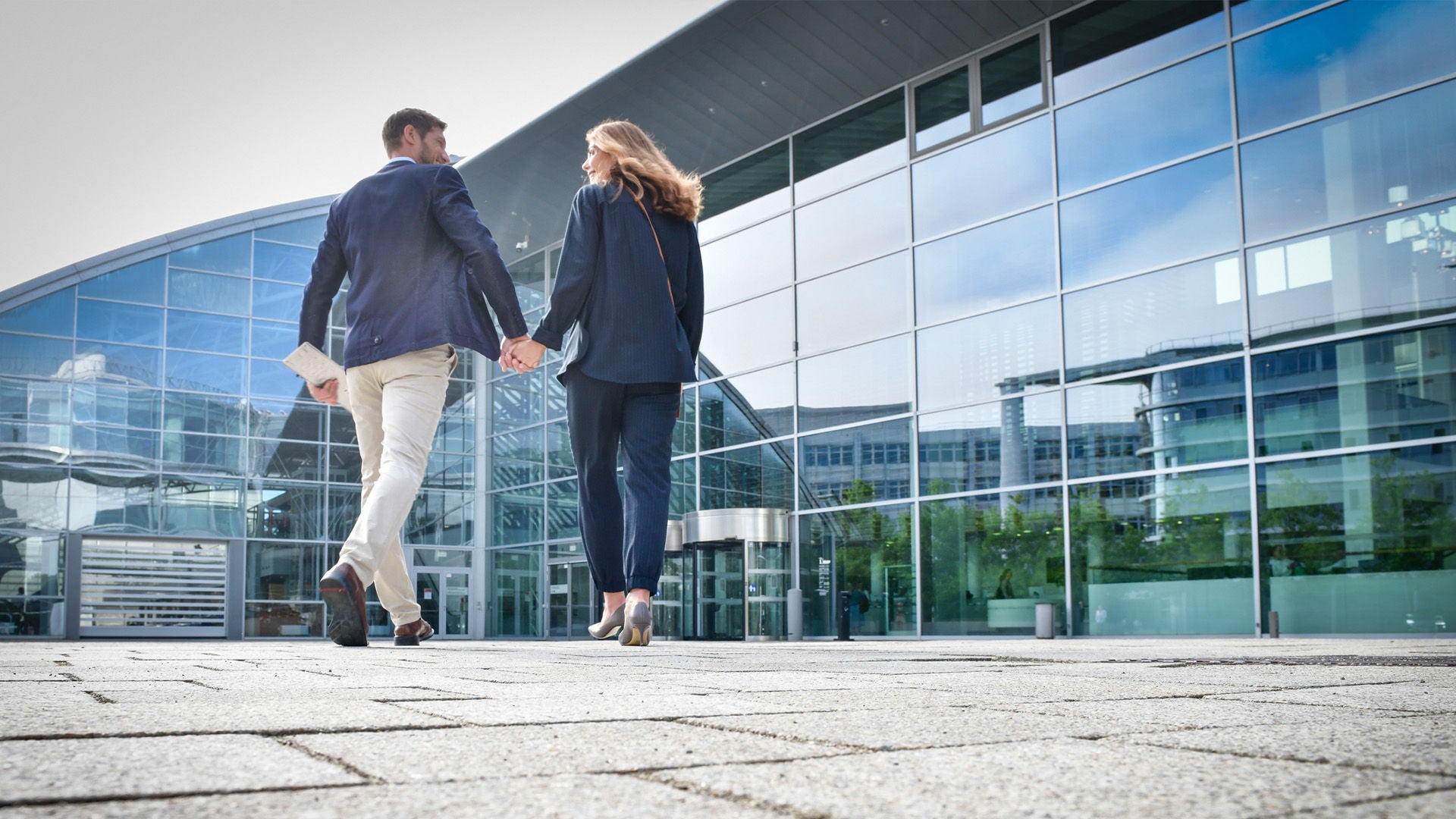 Two people walk hand in hand across a square in front of a modern glass building.