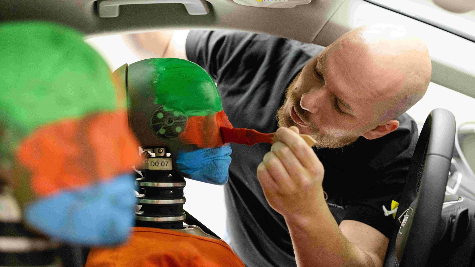 Ein Mann pinselt rote Farbe an den Kopf eines Crashtest-Dummies, der auf dem Fahrersitz eines Testfahrzeugs sitzt.
