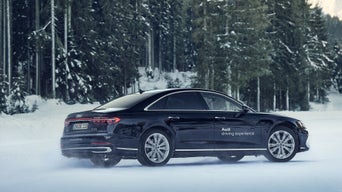 Ein schwarzer Audi A8 50 TDI quattro fährt seitlich über eine Schneefläche, umgeben von verschneitem Wald, Schriftzug „Audi driving experience“ an der Tür.