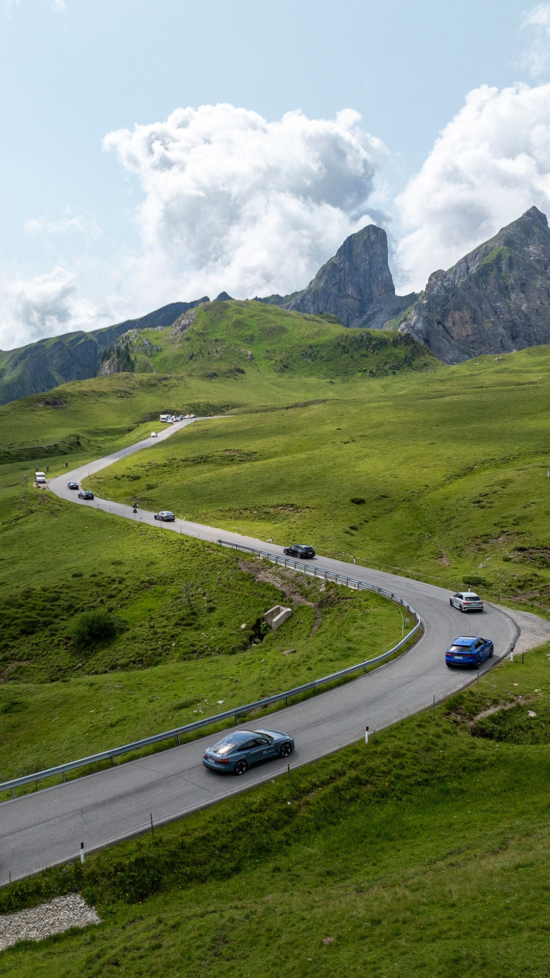 Several Audi vehicles are driving in single file on a winding road through a green, hilly landscape.