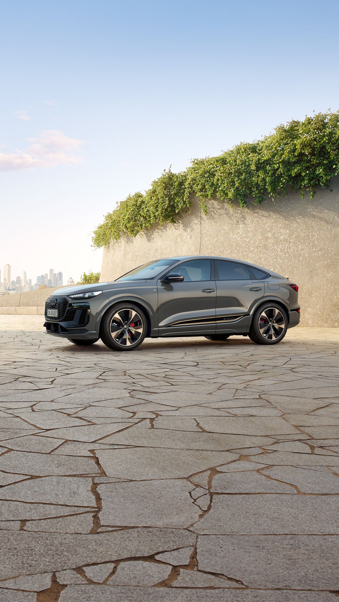 Der Q6 Sportback e-tron steht schräg seitlich vor einer begrünten Steinwand. Im Hintergrund ist die Skyline einer Stadt zu erkennen.