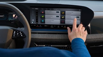 Hand tippt auf großes zentrales Autodisplay mit Fahr-Challenges; daneben Lenkrad und digitale Instrumente sichtbar.