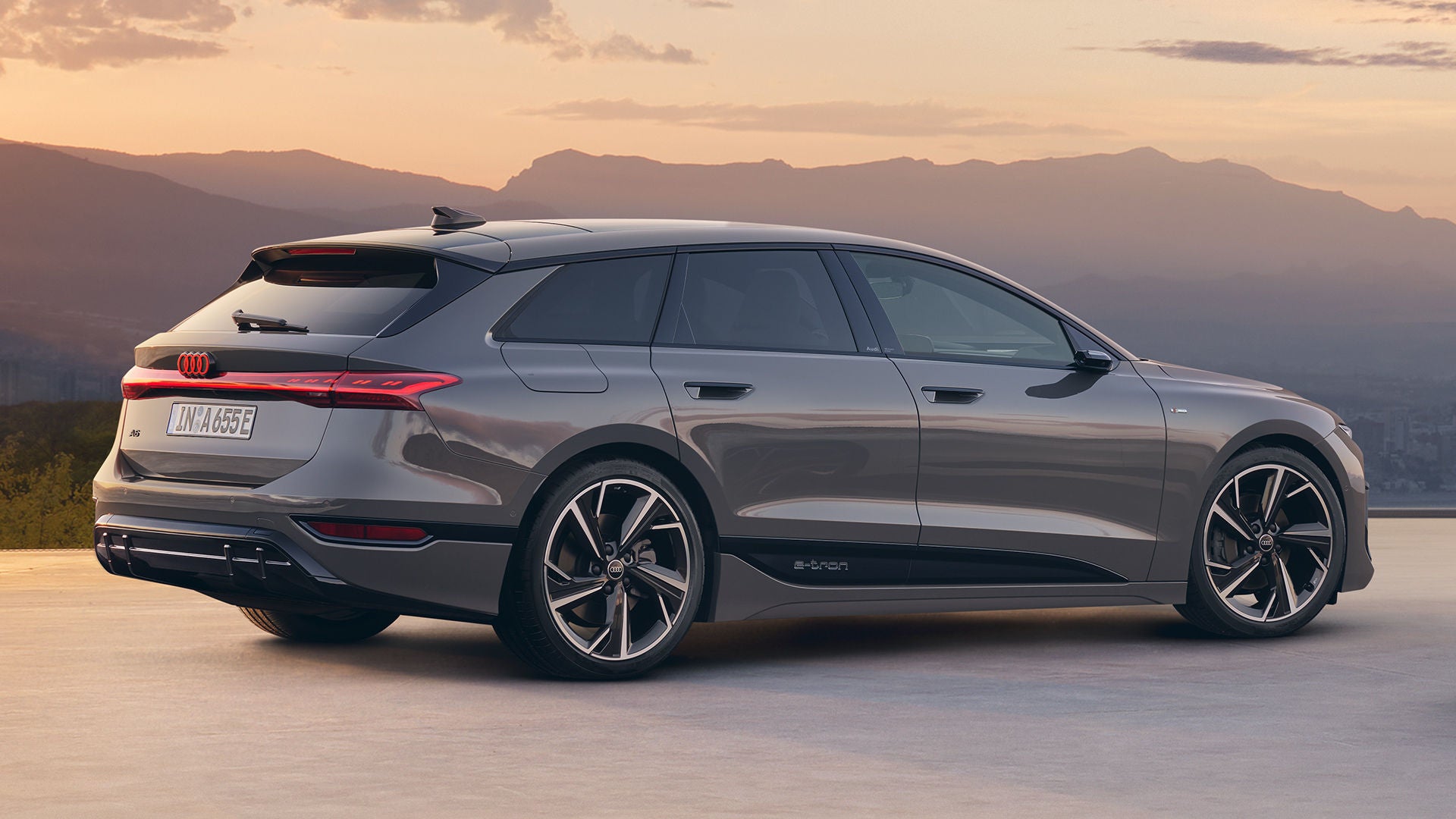 Grauer Audi A6 Avant e-tron in Seitenansicht bei Sonnenuntergang vor Bergsilhouette.