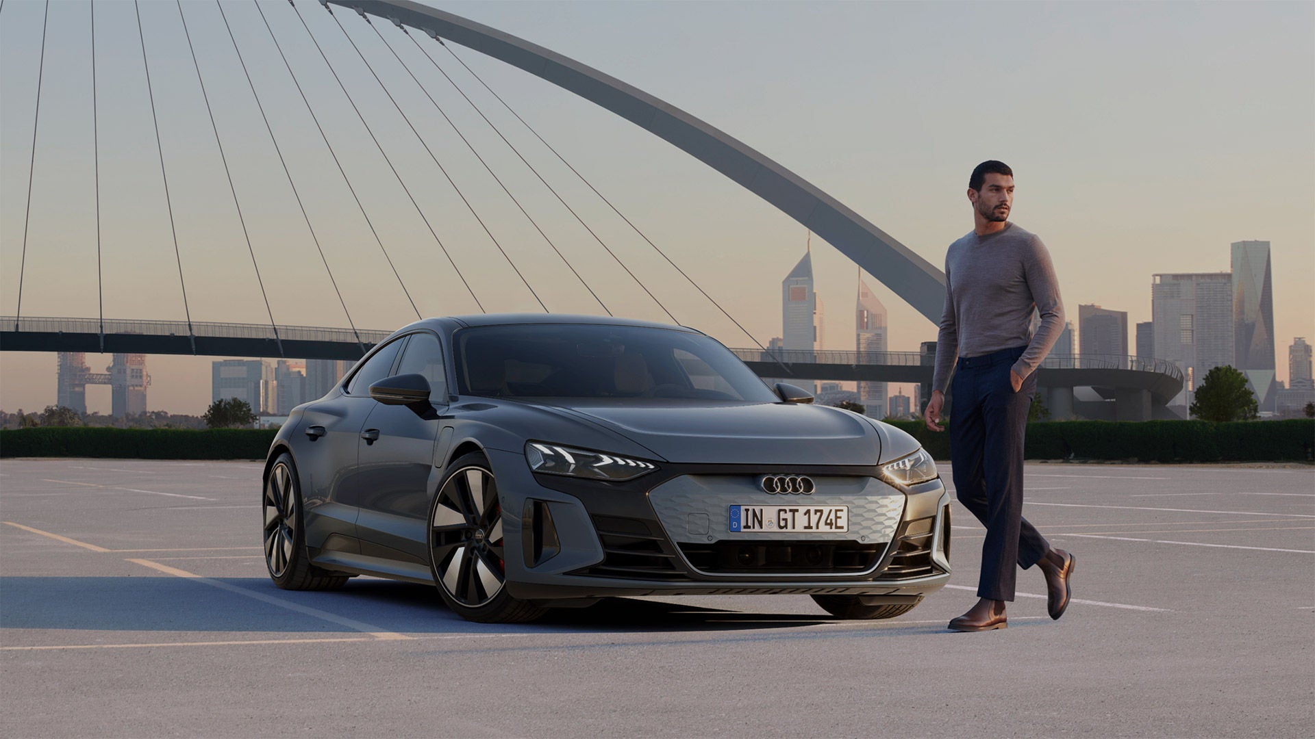 A parked Audi vehicle, a man walking to the right of the vehicle, bridge & cityscape in the distance