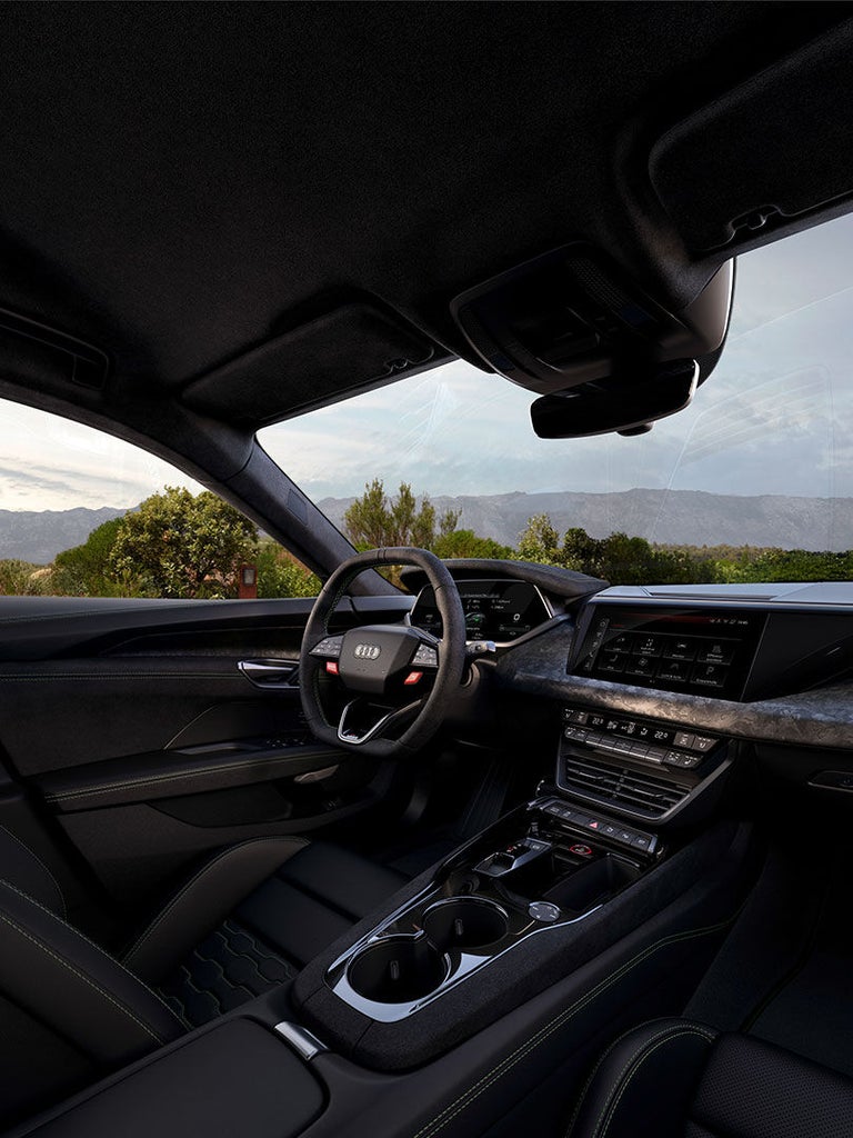 Innenraum eines Audi RS e-tron GT performance mit schwarzem Leder, Lenkrad, Touchscreen und Blick auf grüne Landschaft mit Bergen.
