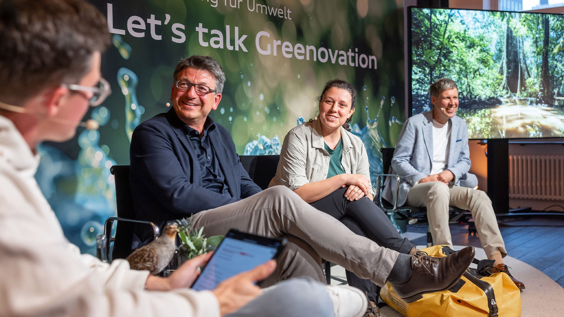Vier Personen sitzen auf einer Bühne und sprechen lächelnd in einer Diskussionsrunde mit dem Titel „Let’s talk Greenovation“.