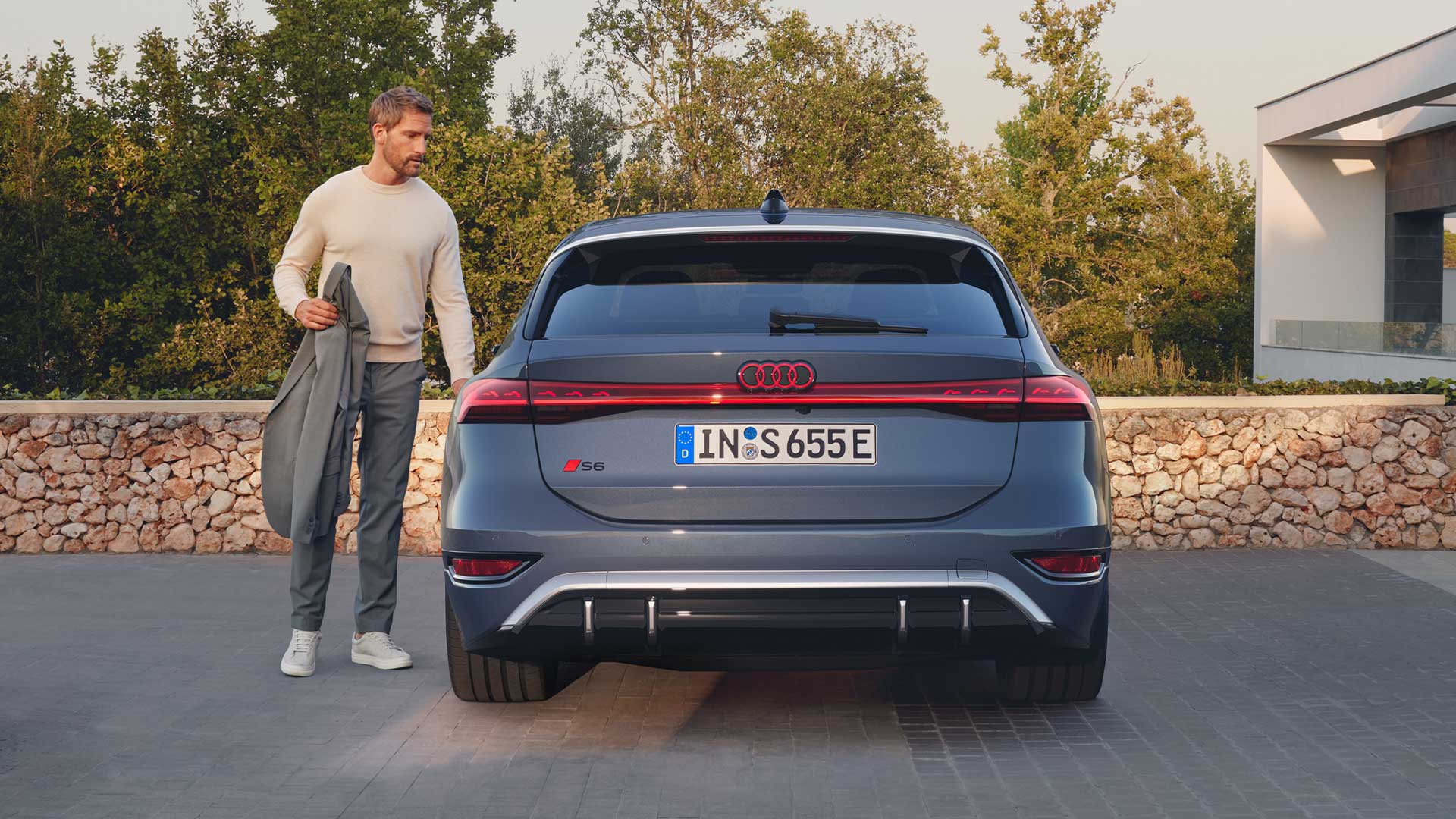 Ein Mann steht neben dem Heck eines geparkten Audi S6 Avant e-tron vor einer Steinmauer mit Bäumen im Hintergrund.
