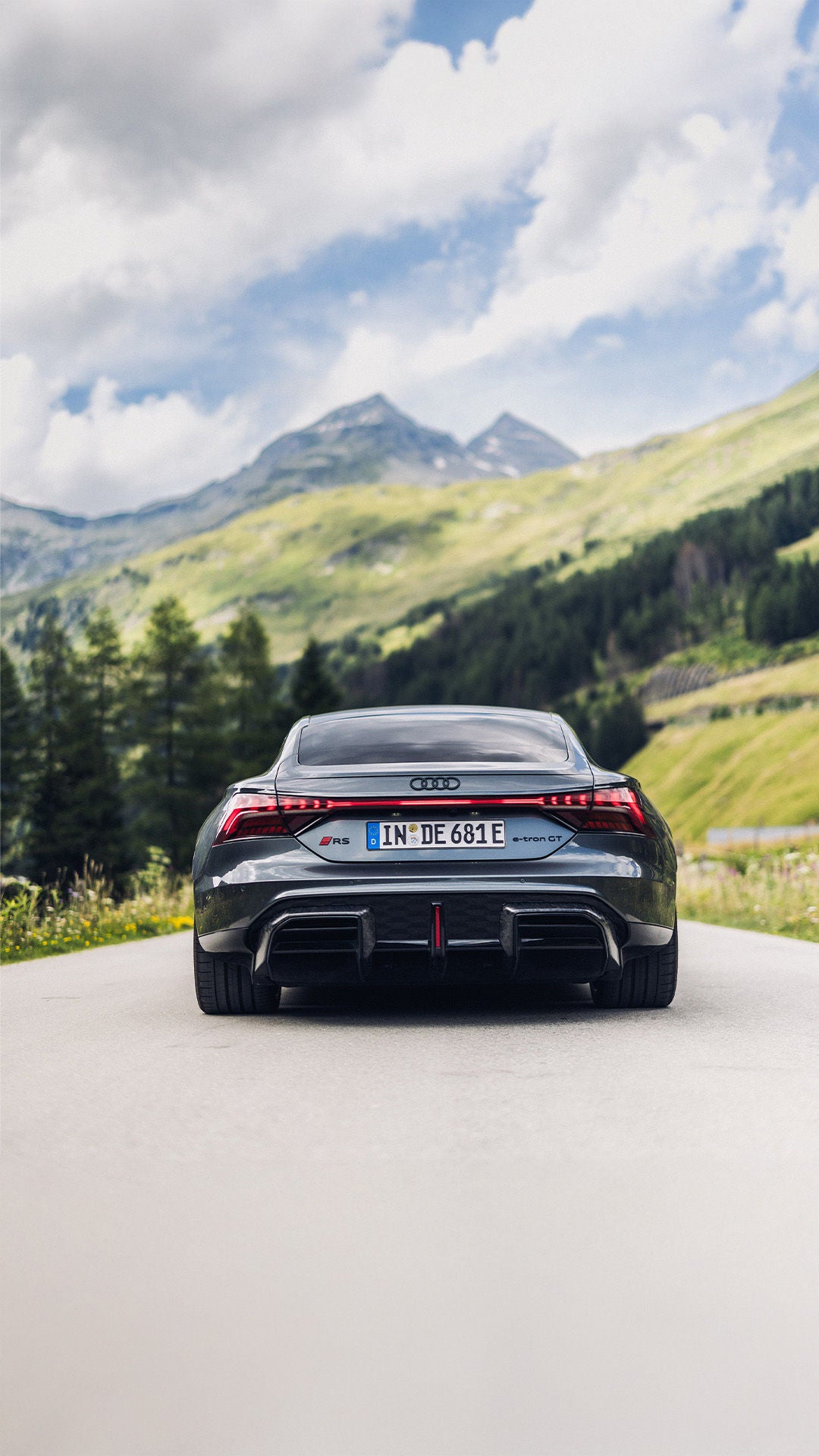Dunkelgrauer Audi RS e-tron GT performance (Heckansicht) fährt auf einer Bergstraße, im Hintergrund grüne Hügel und schneebedeckte Gipfel.