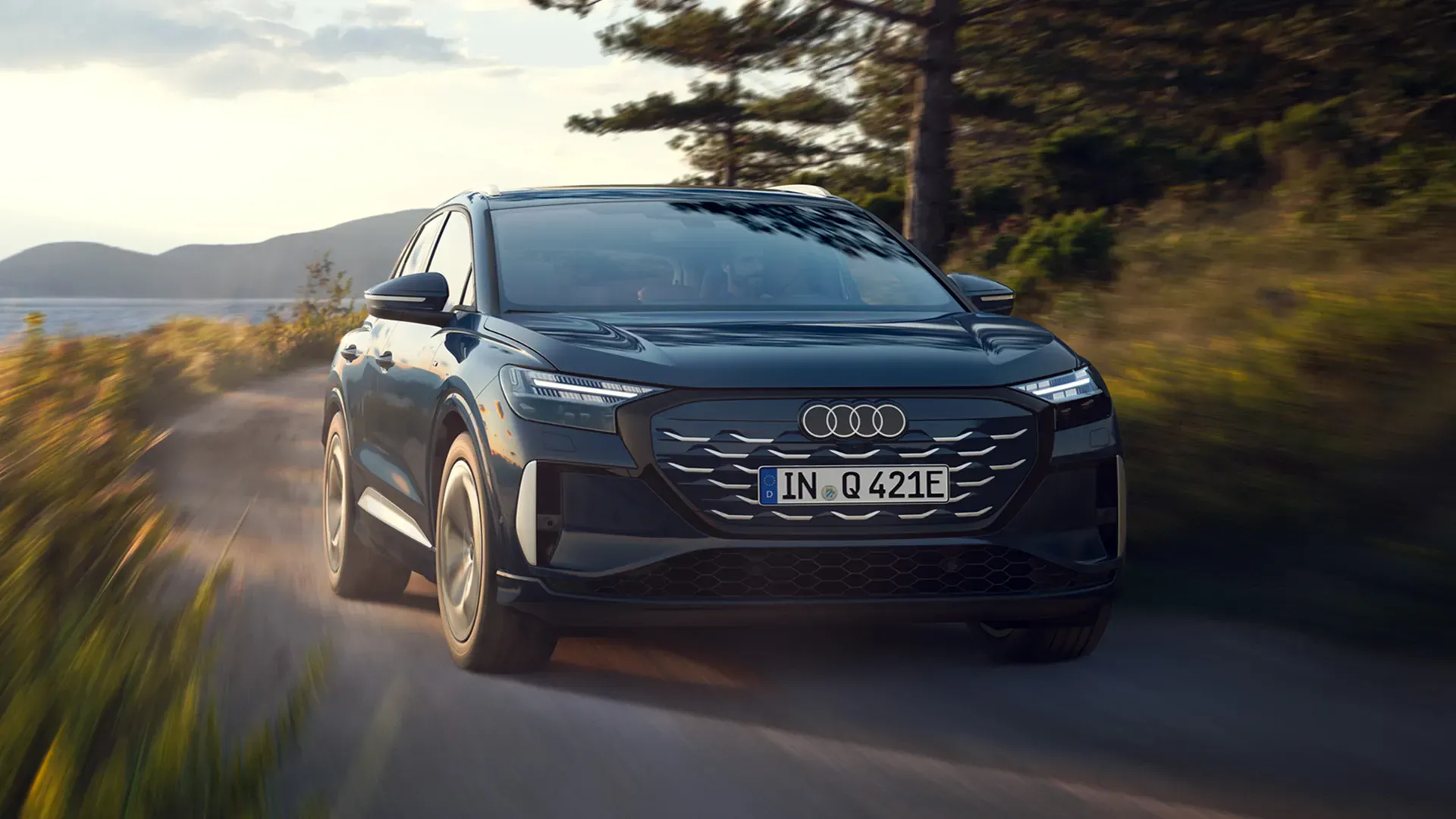 Blue Audi Q4 e-tron EV driving on a scenic coastal road near water and trees