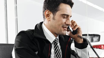 Mann im Anzug und Jacke spricht freundlich mit einem Kunden am Telefon in einem Autohaus.