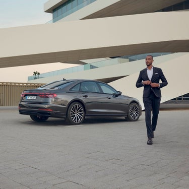 A man in a suit walks near a parked sedan in a modern architectural setting.