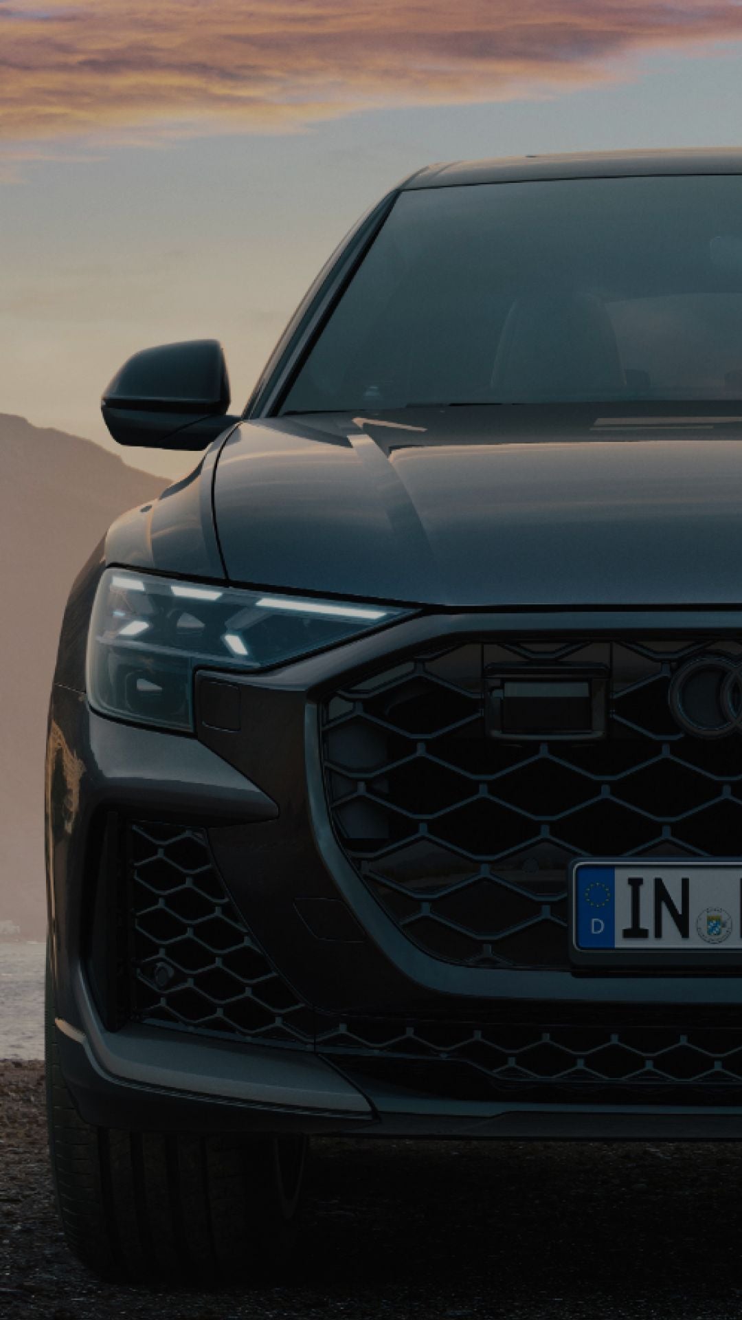 Front of an Audi vehicle, illuminated headlights, cloudy sky in the background in the early evening