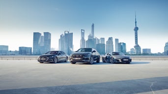 Drei Audi-Modelle nebeneinander vor moderner Skyline mit Fernsehturm und Hochhäusern bei klarem, blauem Himmel.