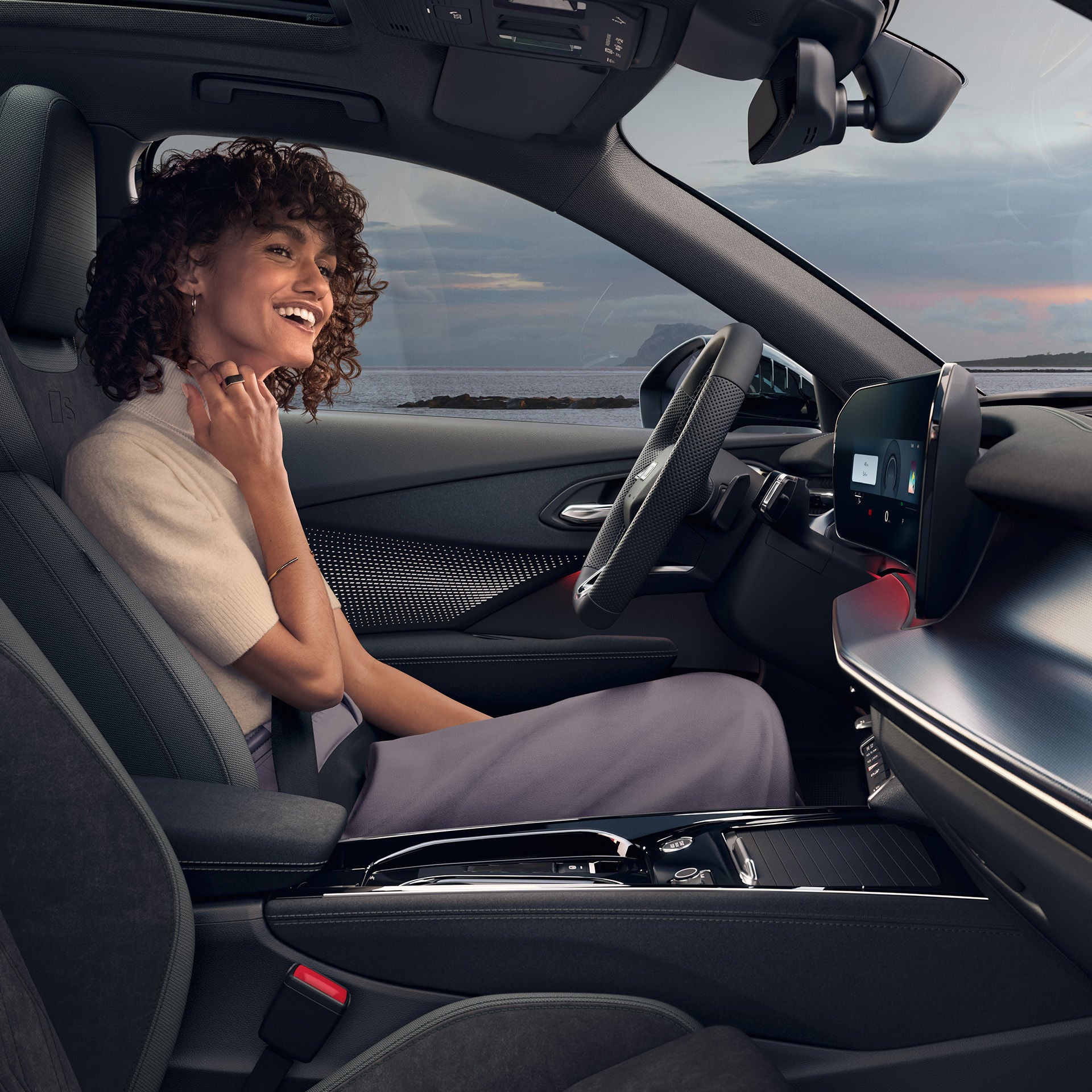 Eine Frau sitzt lächelnd in einem Audi Modell und wird aus der Beifahrerperspektive aufgenommen. Durch die Frontscheibe ist ei Sonnenuntergang zu sehen.