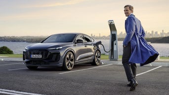 The Q6 Sportback e-tron is parked in a car park and connected to a charging station. A middle-aged man stands in the foreground, with the sea and a city skyline visible in the background.