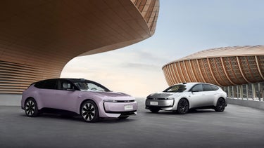 Two Audi E5 Sportbacks in pastel purple and white stand in front of modern architecture.
