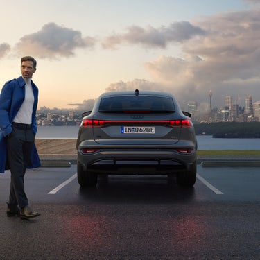 Ein Mann im blauen Mantel steht neben einem geparkten, grauen Audi Q6 Sportback e-tron, im Hintergrund Wasser und Skyline bei Abendstimmung.