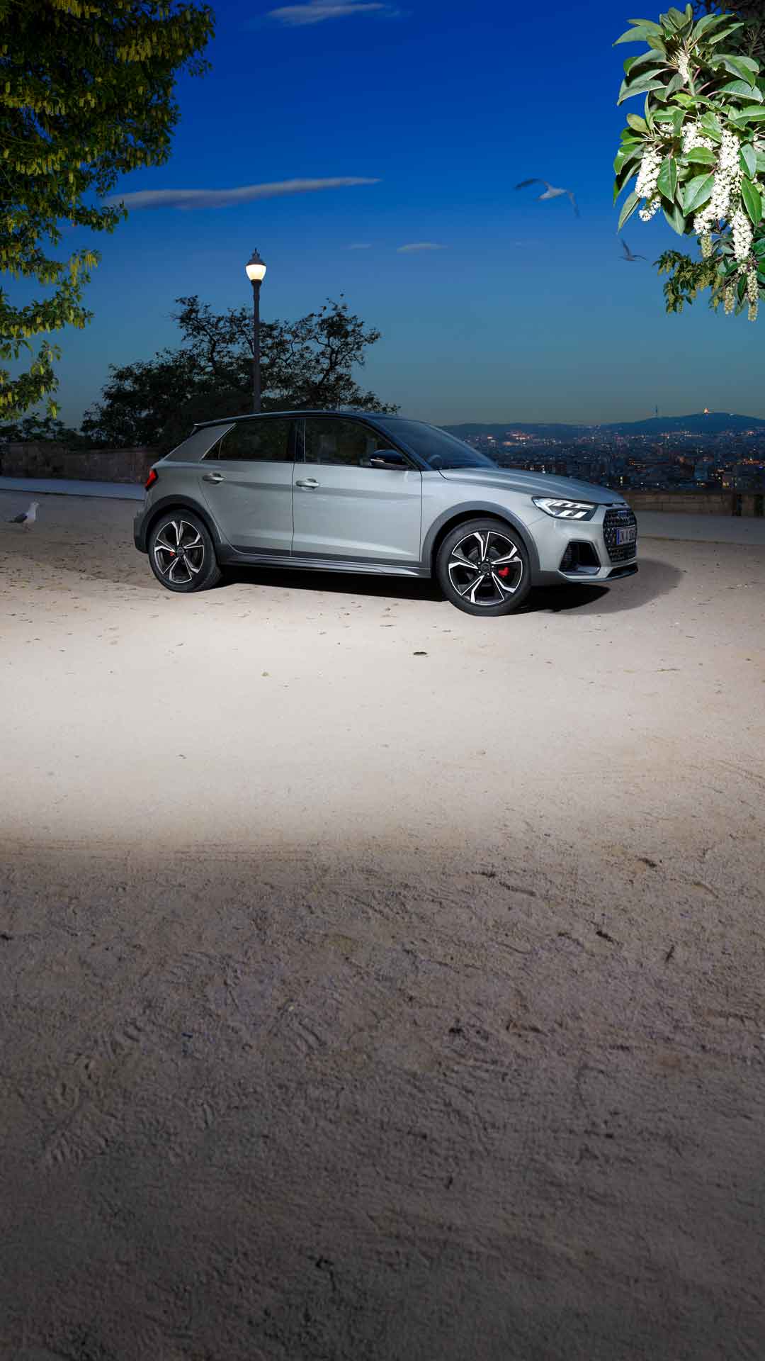 Silberner Audi A1 allstreet im Profil, geparkt auf heller Fläche mit Blick auf beleuchtete Stadt bei Nacht.